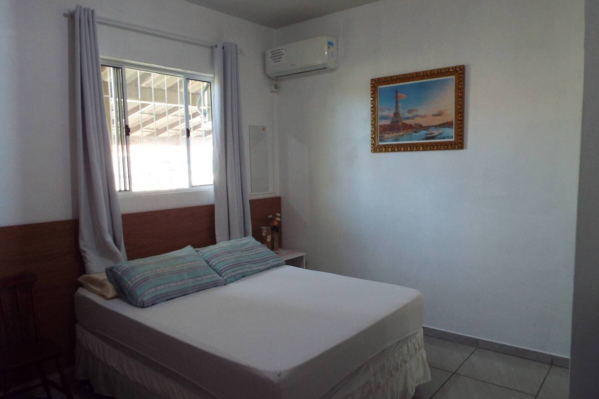 Une chambre à coucher avec un lit, des rideaux, une table de chevet, un tableau encadré et un appareil de climatisation.