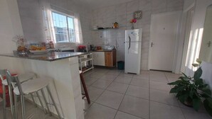 A kitchen with a white refrigerator, a sink, and a window with curtains. There are chairs and a potted plant. - POUSADA DECARLI (Florianopolis)