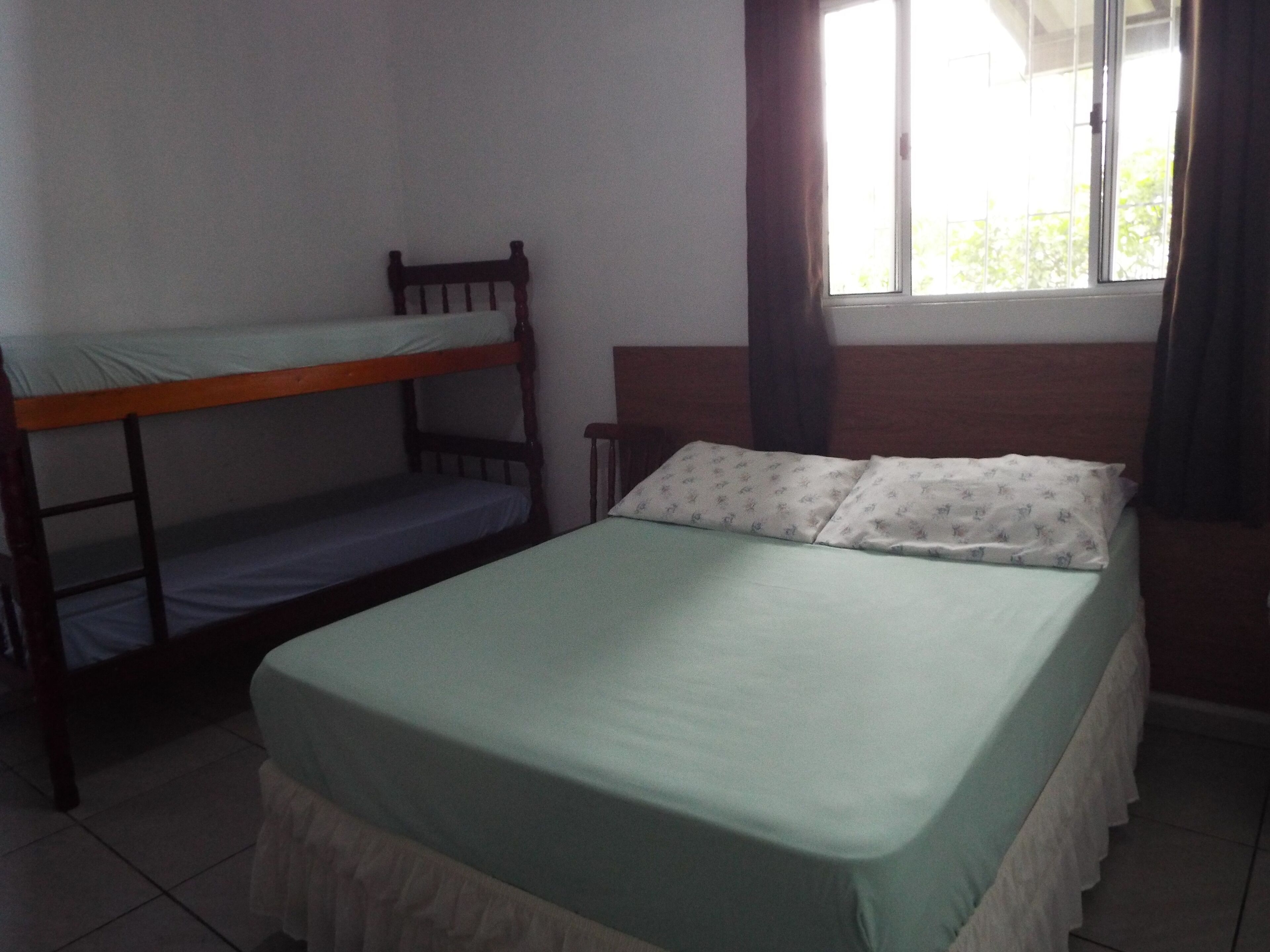 Una stanza con un letto a castello e un letto singolo, entrambi con biancheria da letto verde. Si apre su una finestra con tende, e le pareti sono bianche con un pavimento piastrellato.