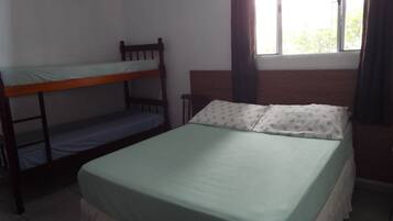 Una stanza con un letto a castello e un letto singolo, entrambi con biancheria da letto verde. Si apre su una finestra con tende, e le pareti sono bianche con un pavimento piastrellato.