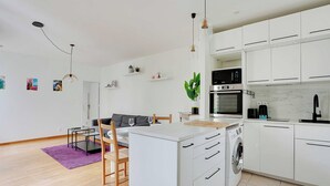 Une cuisine moderne avec des meubles blancs, un lave-linge et un coin repas agrémenté d’un tapis violet et de chaises en bois.