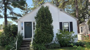 Una casa pequeña de color blanco con una puerta verde oscuro, un porche y un alto árbol de hoja perenne al frente.