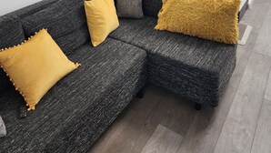 Ett modernt vardagsrum med en hörnsoffa, ett litet köksutrymme med mikrovågsugn och vattenkokare samt en stor platt-TV som är monterad ovanför köksbänken.