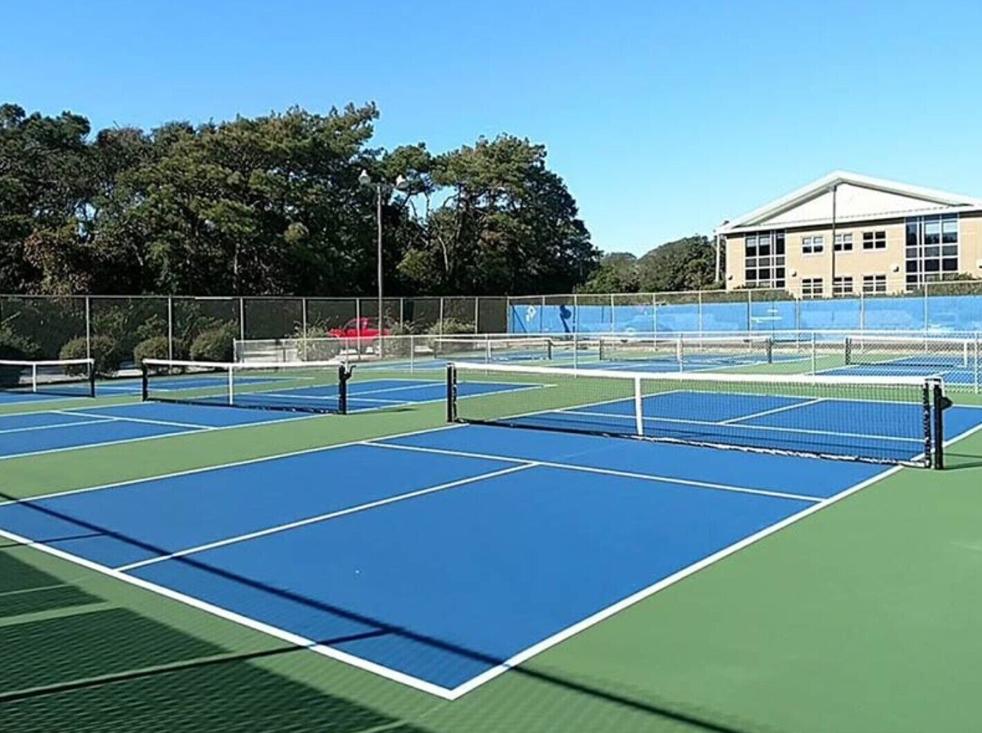 An outdoor tennis court with multiple courts, surrounded by a fence and trees.