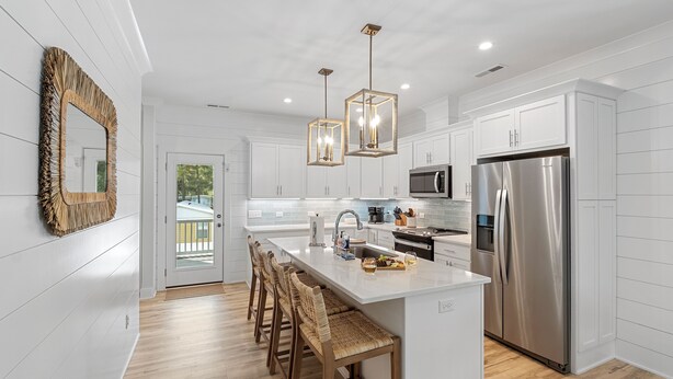 A modern kitchen with white cabinetry, a central island with bar stools, stainless steel appliances, and pendant lighting. - Modern, Luxury Beach House with Private Pool and Hot Tub - Sleeps 12 (Oak Island)
