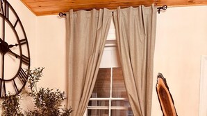 A cozy room with a wooden ceiling, a large wall clock, a window with beige curtains, two wooden chairs, a small table with a vase, and a potted plant. - The Aviator: Serene Ambience & Stylish Comfort (Madison)