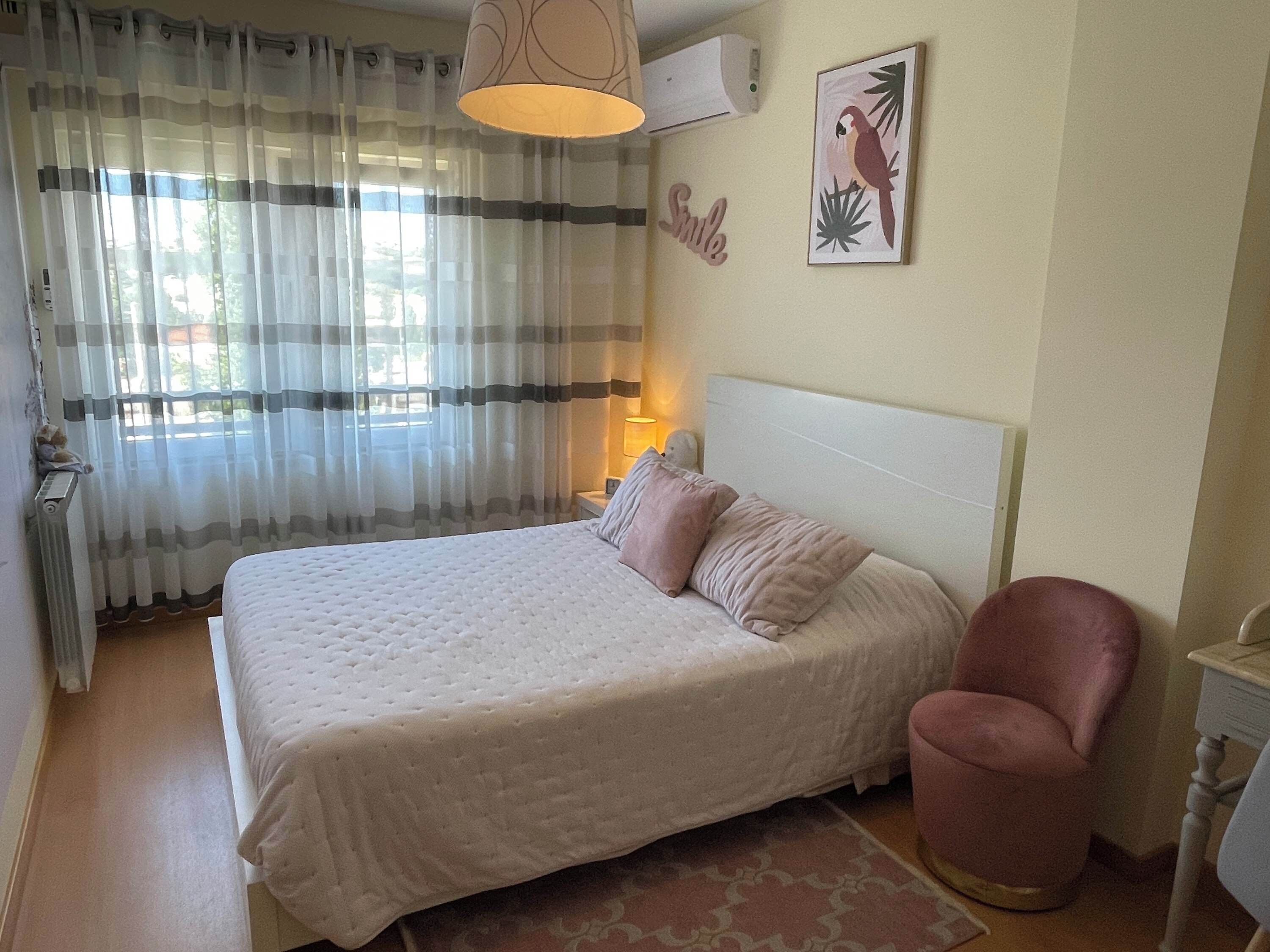 A neatly made bed with a white headboard, a pink chair, and a radiator on the left.