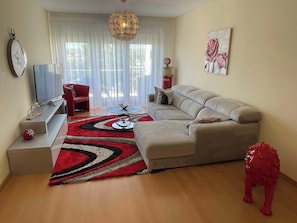 A living room with a sofa, a coffee table, a red decorative dog statue, and a large window with sheer curtains.