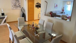Un área de comedor moderna con una mesa de vidrio y sillas blancas, un espejo decorativo y vistas de una sala de estar con un sofá y una mesa de comedor.