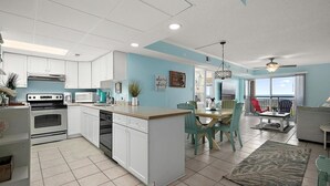A modern kitchen with white cabinets, a central island, and a dining area with teal chairs. - Surfcaster 103 (Ocean City)