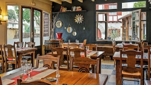 A restaurant interior with wooden tables and chairs, a black wall with decorative mirrors, and large windows offering a view of the outside. - Hôtel Spa Mendi Alde (Ossès)
