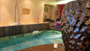 A wooden table with a decorative vase and a small candle. In the background, there is a pool area with a tiled floor and a wall-mounted clock. - Hôtel Spa Mendi Alde (Ossès)
