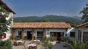 A building with a tiled roof, outdoor seating area, and a view of mountains in the background. - Hôtel Spa Mendi Alde (Ossès)
