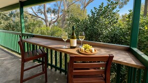 Uma mesa de madeira com uma garrafa de vinho branco, duas taças e um prato de queijo e uvas numa varanda com grades verdes e vista para árvores.