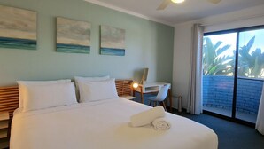 A hotel room with a large bed, a desk, and a ceiling fan. There are paintings on the wall and a view of palm trees through the window. - Coastal Scarborough Beach • Chic • Pool View • 2BR (Scarborough)