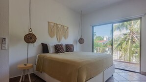 A bedroom with a bed, a small bedside table, a macrame wall hanging, and a view of palm trees through a large window. - Spacious 2 bdrm + A/C, 2 bdrm + A/C in Playacar (Playa del Carmen)