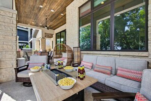 Une terrasse extérieure avec une longue table, des chaises en osier, un canapé et de grandes fenêtres.