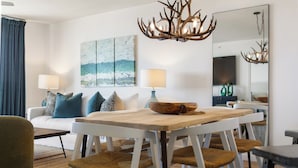A modern living room with a wooden dining table, white chairs, a sofa with blue cushions, and a large painting on the wall.