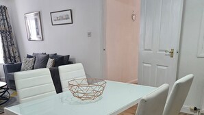 Un comedor con una mesa y sillas blancas, un cuenco geométrico sobre la mesa, y un sofá con cojines al fondo.