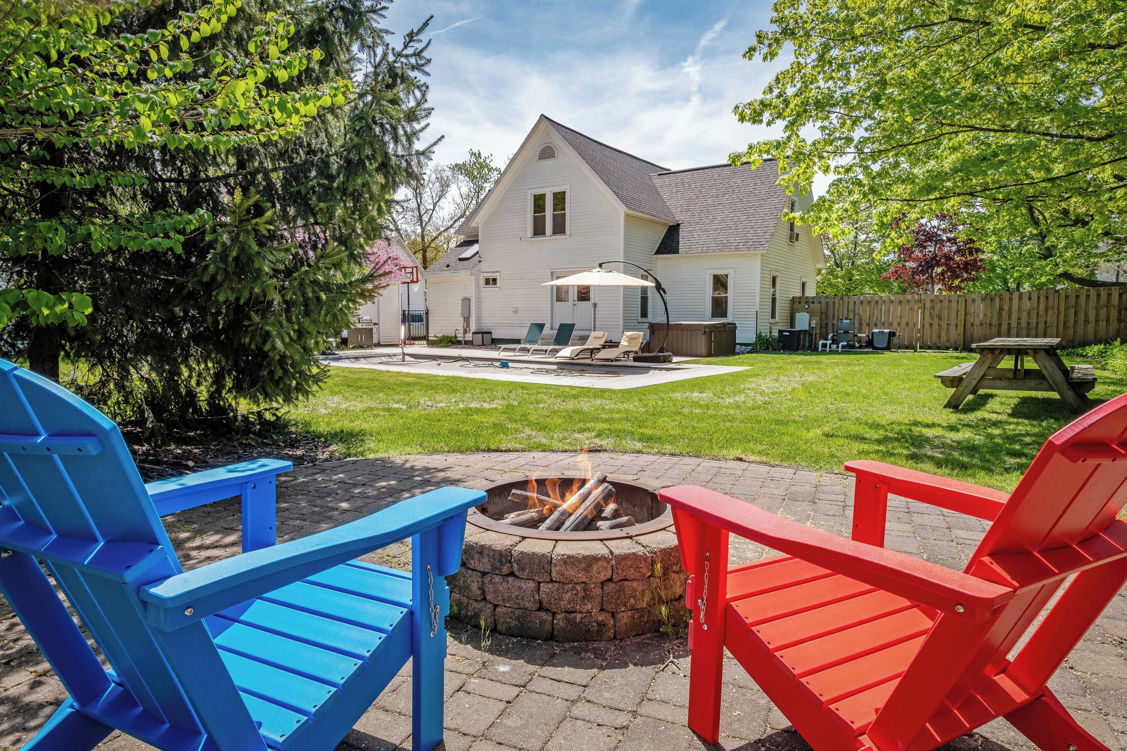 Un patio trasero con un brasero, dos sillas Adirondack, una mesa de madera y una casa al fondo.
