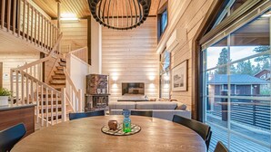 A wooden interior with a round dining table, a staircase, and a large window offering a view of the outside.