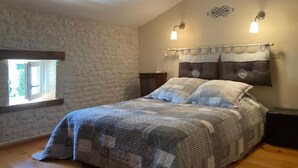 A bedroom with a stone wall, a bed with a patterned bedspread, two wall-mounted lamps, and a window with a view of greenery. - GITE DES VIGNES (Archiac)