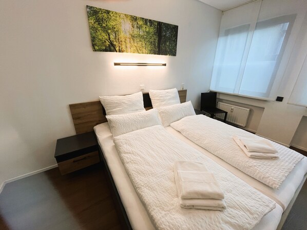 A neatly made bed with white linens and towels, a nightstand with a lamp, a painting of a forest, and a window with blinds.