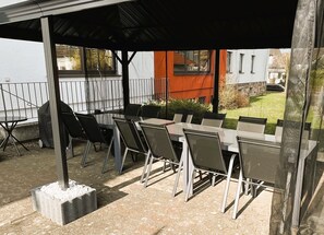 An outdoor patio with a covered table and chairs, a grill, and a view of a building with a red roof.