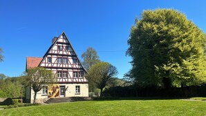 Une maison traditionnelle à colombages, dotée d’un toit en pente, d’un escalier apparent menant à l’entrée et d’une pelouse bien entretenue parsemée de fleurs.