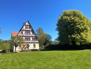 A traditional half-timbered house with a sloped roof, a visible staircase leading to the entrance, and a well-maintained lawn with scattered flowers.