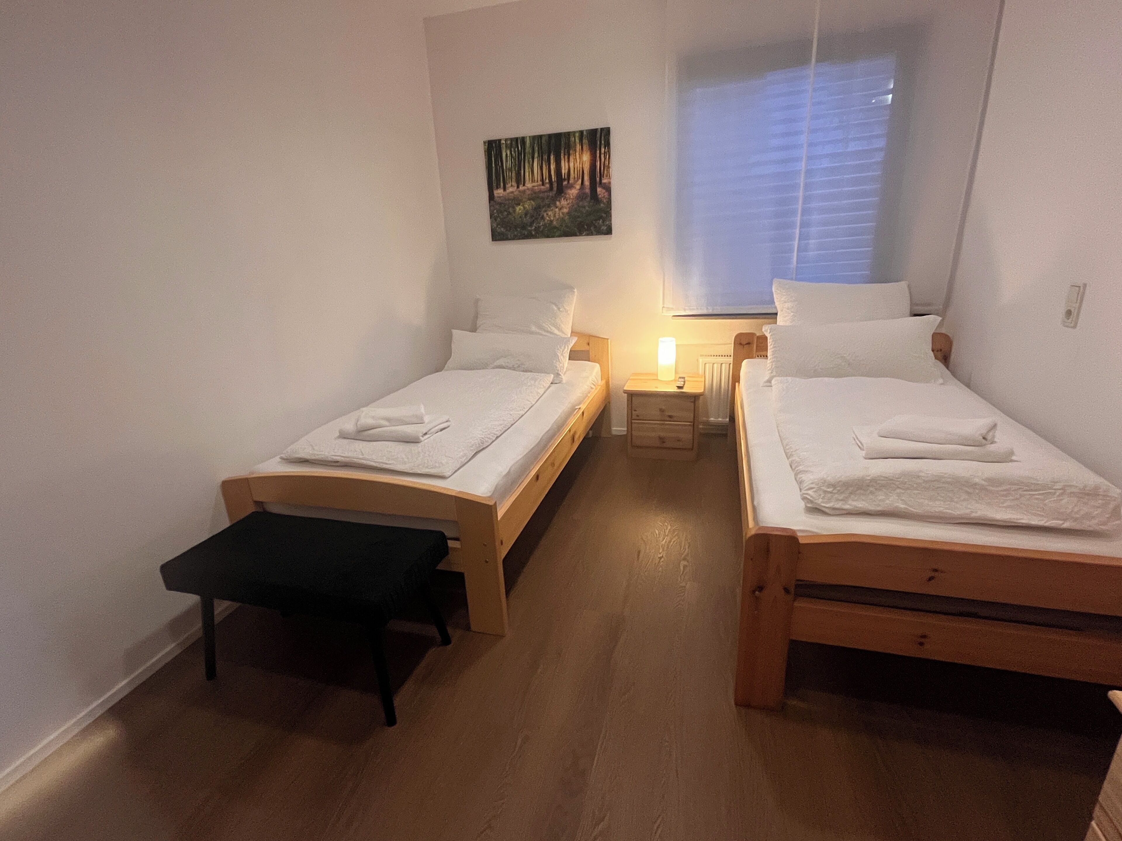 A small, cozy room with two single beds, a bench, a nightstand with a lamp, and a painting on the wall.