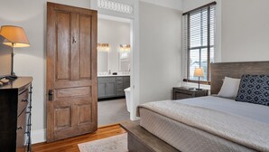 Une chambre avec une porte en bois, une table de chevet avec une lampe, un lit avec une literie à motifs et une fenêtre avec des stores.
