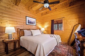 A wooden cabin bedroom with a bed, two bedside lamps, a ceiling fan, a framed picture on the wall, and a window with blinds. - Free Dollywood Tix/Pool/Modern/Luxurious/Secluded (Sevierville)