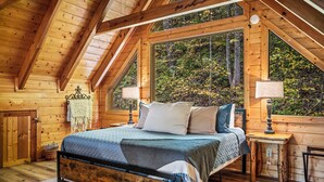 Een slaapkamer in een houten hutje met een bed, twee nachtkastjes en een groot raam met uitzicht op een bos.