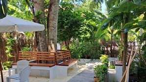 A garden patio with a concrete pathway, white plastic chairs, wooden benches, and a white umbrella. - Shams Thoddoo (Thoddoo)