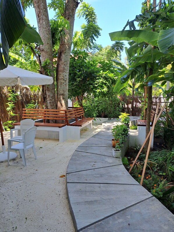 A garden patio with a concrete pathway, white plastic chairs, wooden benches, and a white umbrella.