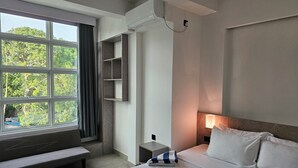 A hotel room with a large window, a bed with pillows, a bedside table, and a bench. - Shams Thoddoo (Thoddoo)