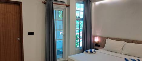 A hotel room with a bed, a bedside table with a lamp, a window with curtains, and an air conditioning unit.