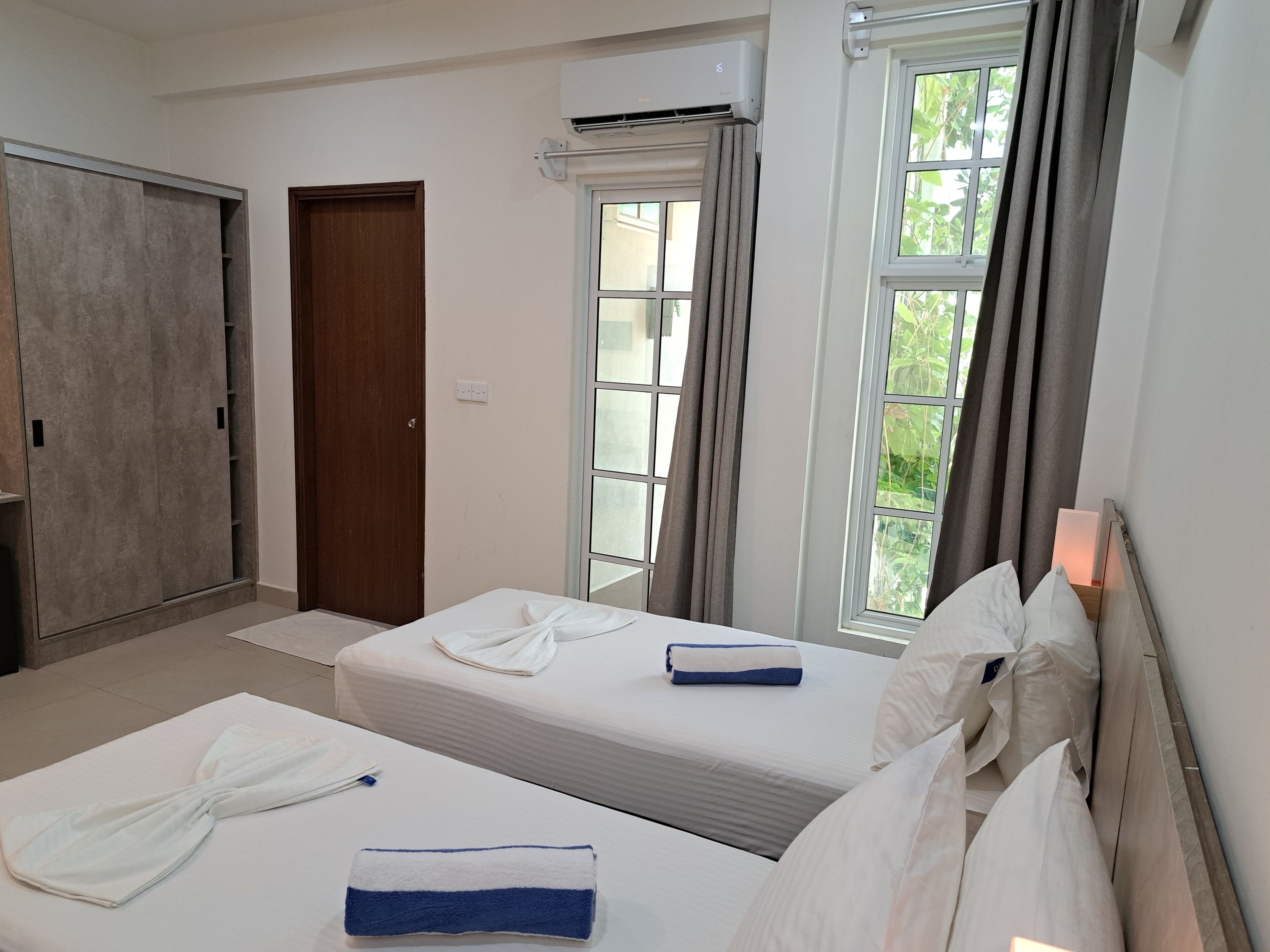 A hotel room with two beds, a wardrobe, an air conditioning unit, and large windows with curtains.