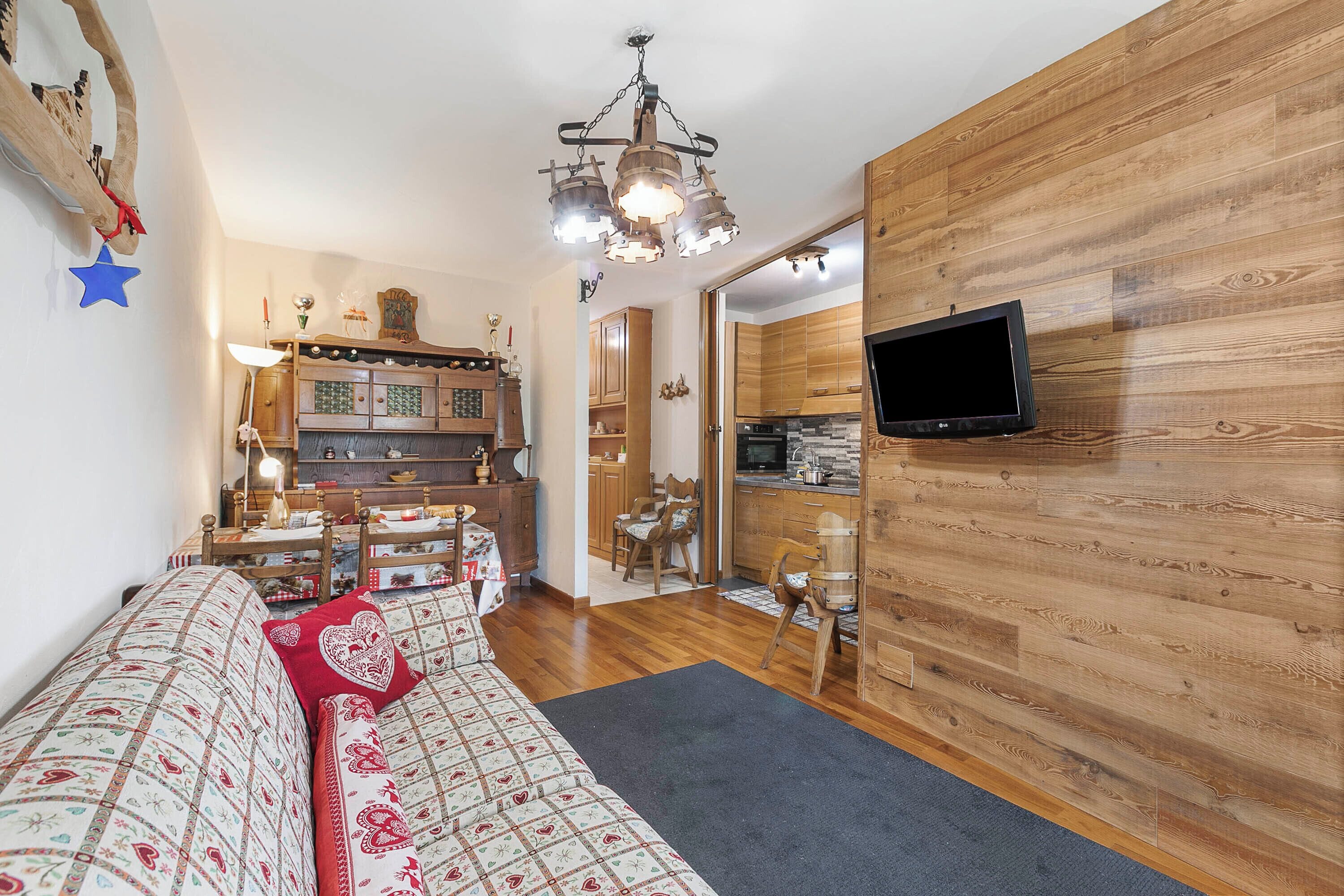 A cozy living room with a sofa, a television mounted on a wooden wall, and a kitchen in the background.