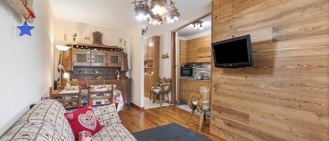 A cozy living room with a sofa, a television mounted on a wooden wall, and a kitchen in the background.