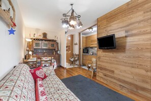 Un salon chaleureux avec un canapé, un téléviseur fixé à un mur en bois, et une cuisine en arrière-plan.
