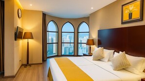 Ein Hotelzimmer mit einem großen Bett, zwei Nachttischlampen, einer Uhr und Blick auf die Stadt durch bogenförmige Fenster.