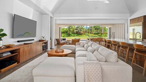 Un salón moderno con un amplio sofá modular, un televisor de pantalla plana, muebles de madera y vistas a un jardín exuberante.