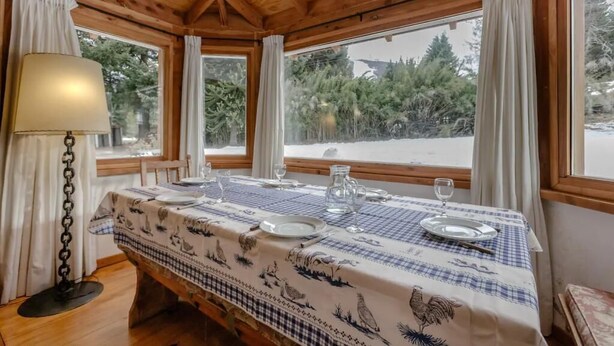 A dining area with a table set for two, a lamp, and a view of trees and snow outside. - Mountain cabin with large garden and lots of sun. (San Carlos de Bariloche)