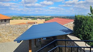 A view from a balcony with a stone wall, a red-roofed building, and a blue-roofed structure with a solar panel. - Winegrower's house, private courtyard, 4 people/2 beds (Prompsat)