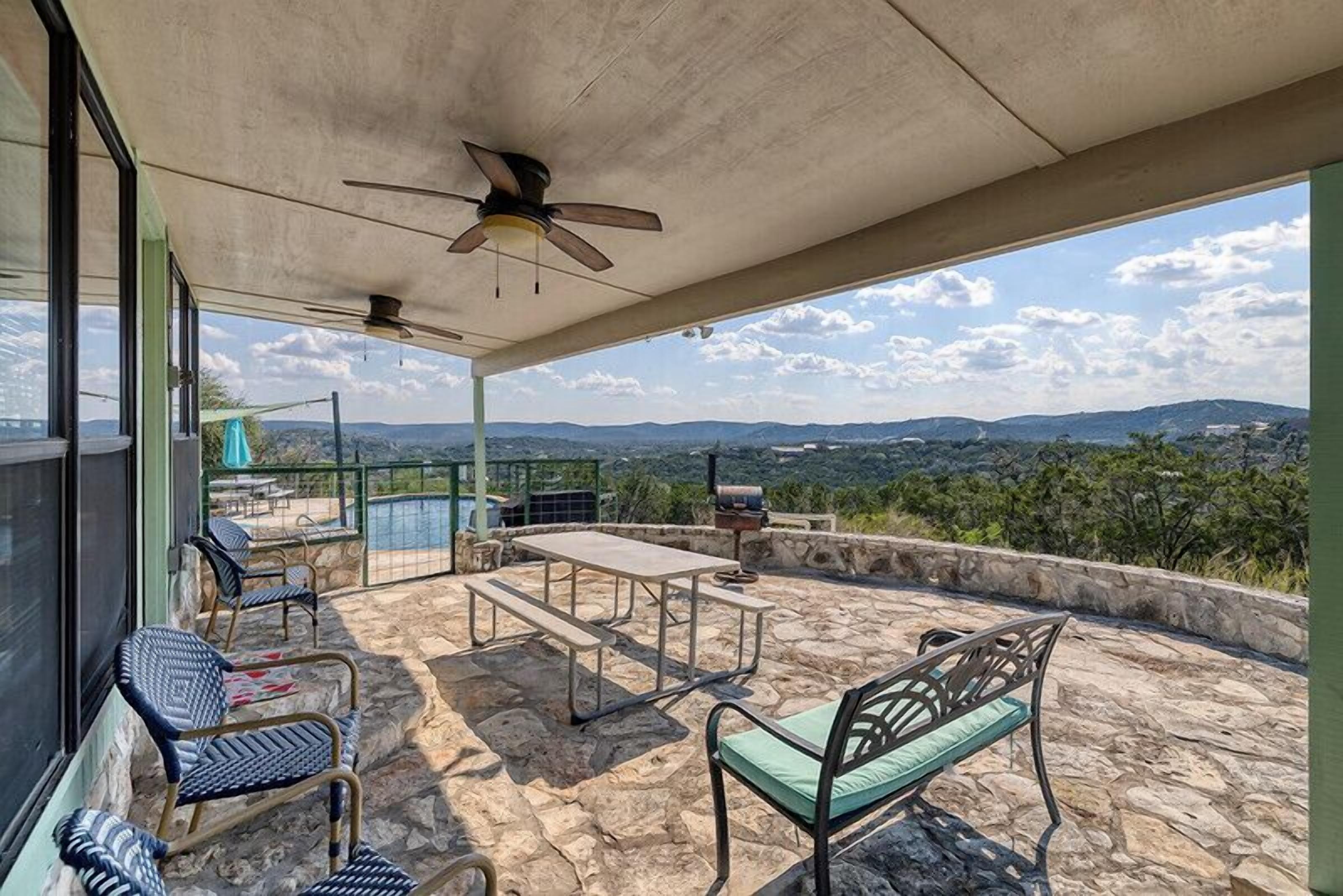 A covered outdoor area with stone flooring, a ceiling fan, a table, chairs, and a view of the landscape.