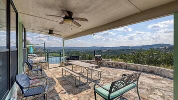A covered outdoor area with stone flooring, a ceiling fan, a table, chairs, and a view of the landscape.