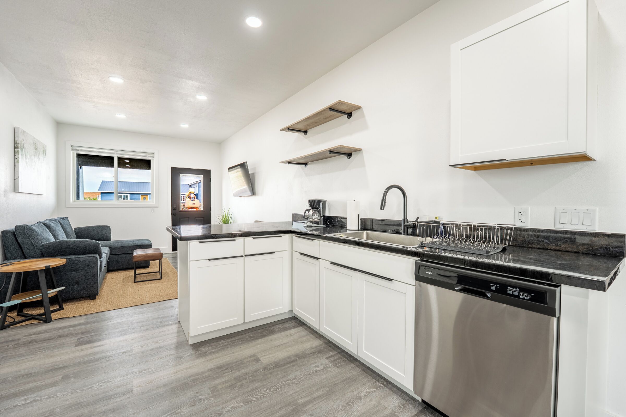 A modern kitchen with white cabinets, a stainless steel dishwasher, and a black countertop. There is a gray sofa and a small wooden table in the adjacent living area.