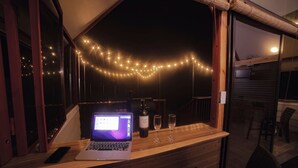 Een laptop op een houten toonbank met twee wijnglazen en een fles wijn, sfeerverlichting erboven en een uitzicht op buiten.